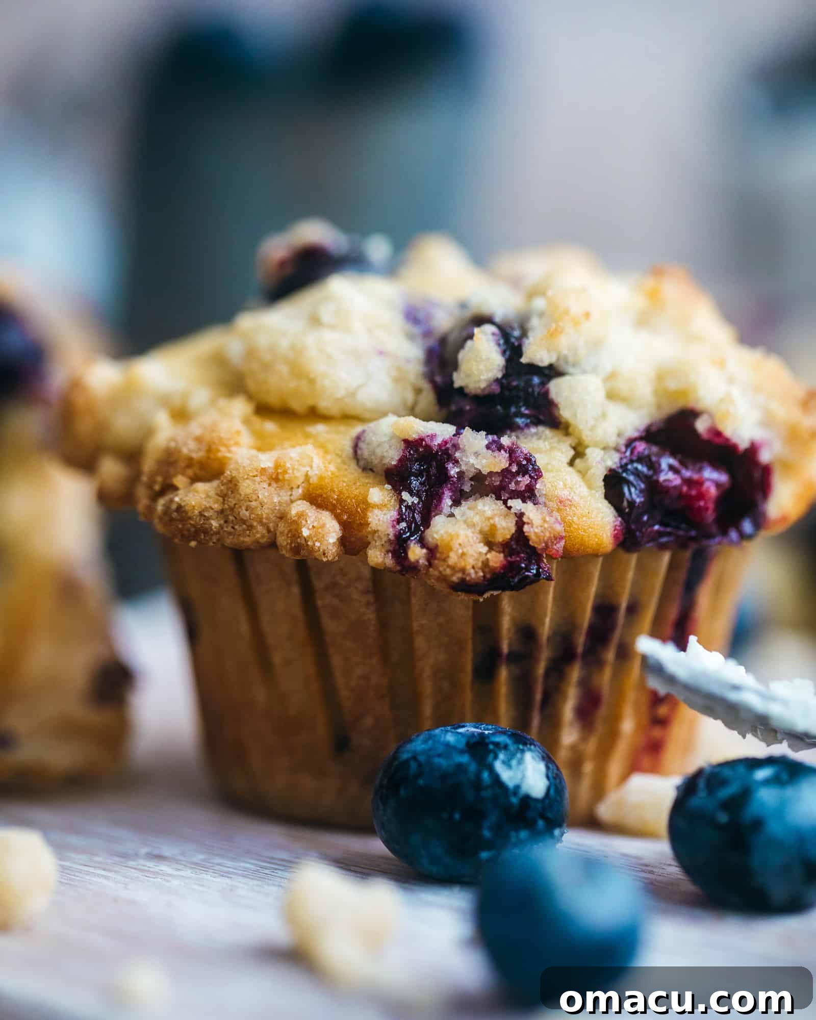 Close-up of a single blueberry crumb cake muffin, with a scattering of fresh blueberries in the foreground