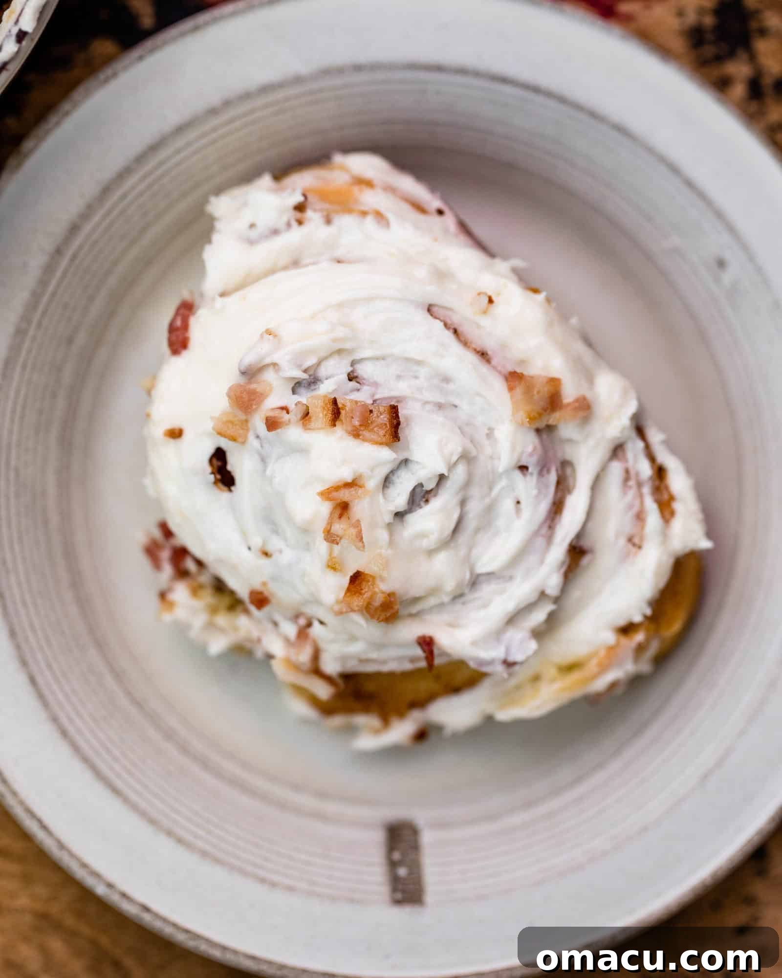 A single Maple Bacon Cinnamon Roll on a white plate, generously topped with maple icing and sprinkled with more crispy bacon bits.