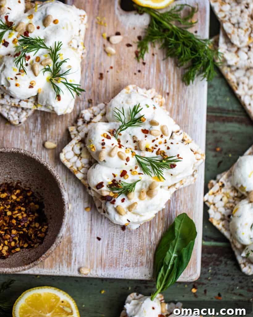 A vibrant display of roasted garlic labneh on a wooden cutting board, garnished with lemon wedges, red pepper flakes, and an assortment of fresh herbs.