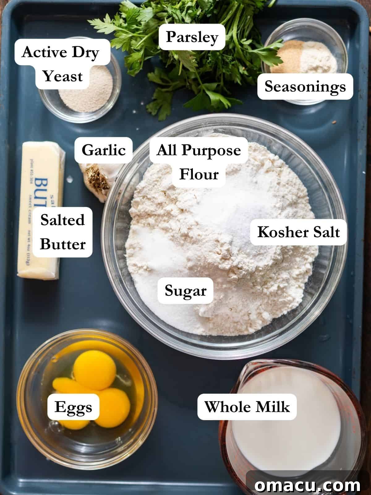 A flat lay of fresh ingredients arranged on a baking sheet to make garlic butter rolls: flour, sugar, salt, garlic powder, onion powder, butter, fresh parsley, eggs, egg yolks, whole milk, and active dry yeast.