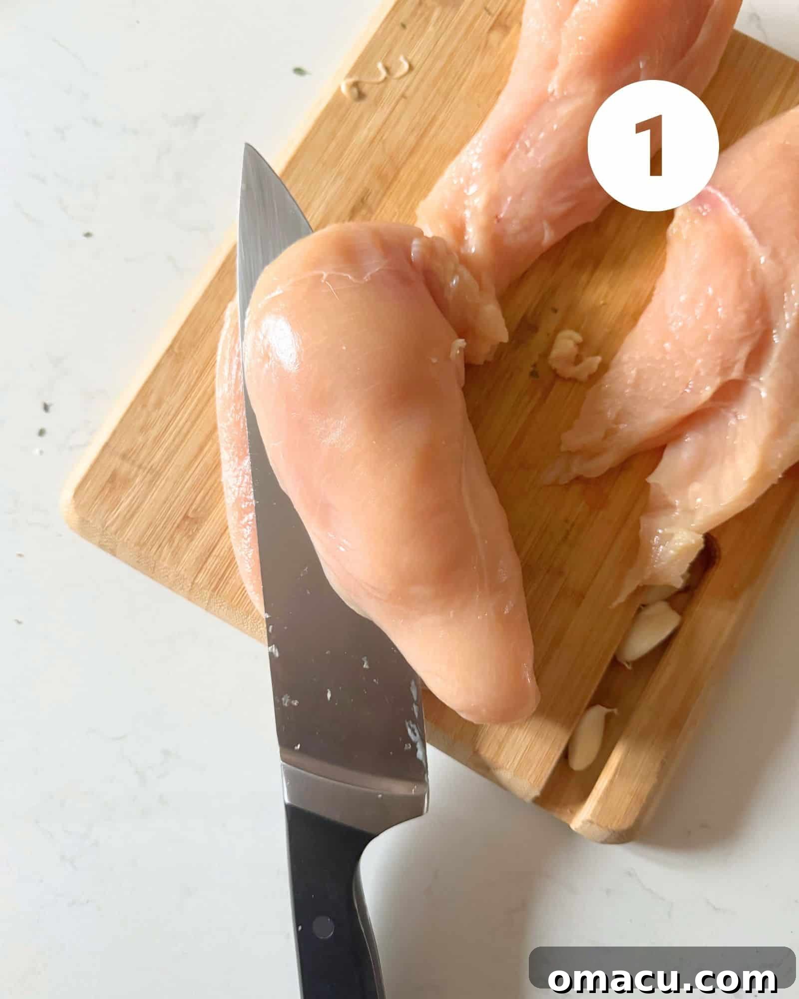 A sharp knife precisely cutting a boneless chicken breast in half lengthwise on a cutting board, preparing it for cooking.