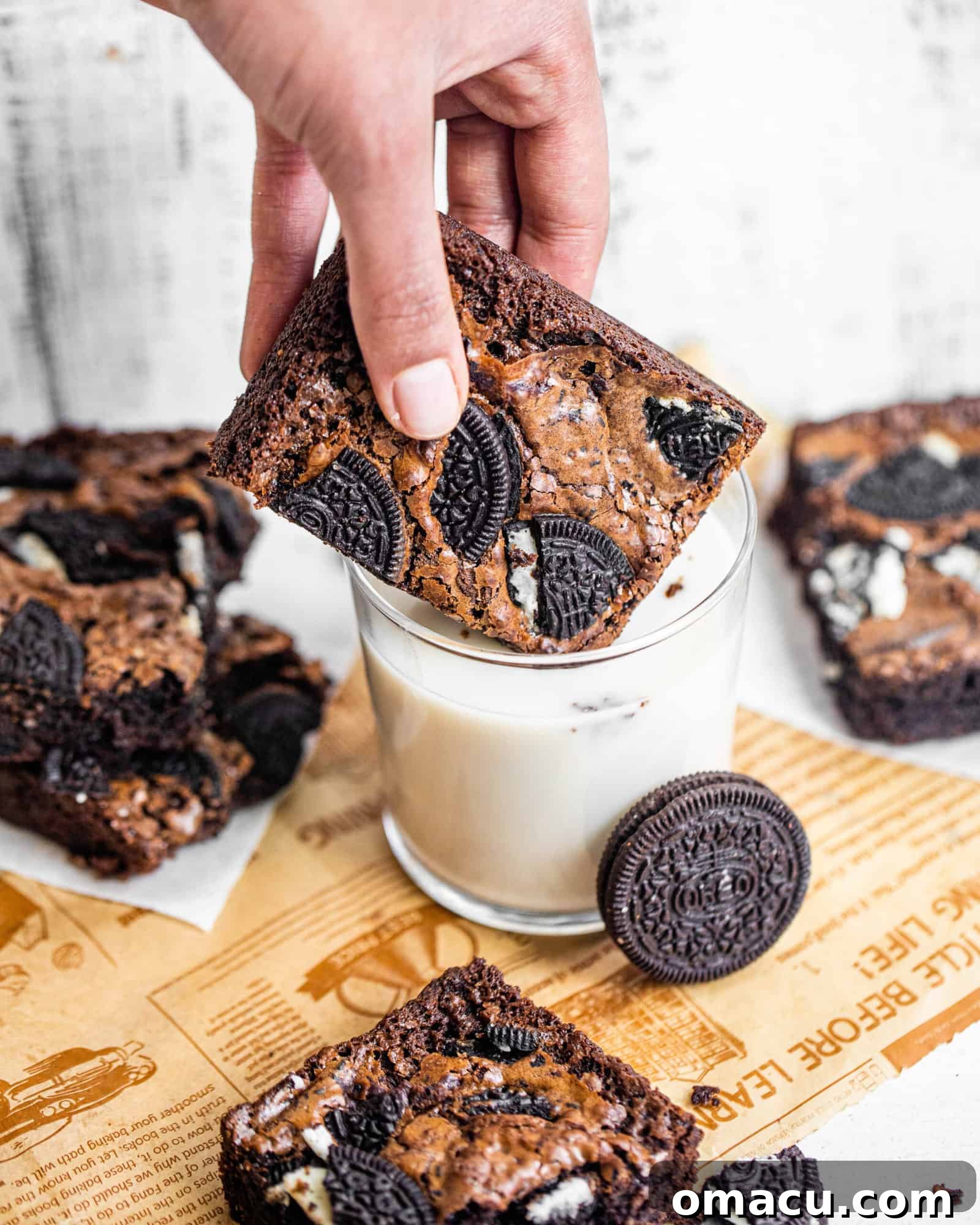 A hand gracefully dipping a fudgy Oreo brownie into a glass of cold milk, highlighting its rich, dark color and the crunchy cookie bits.