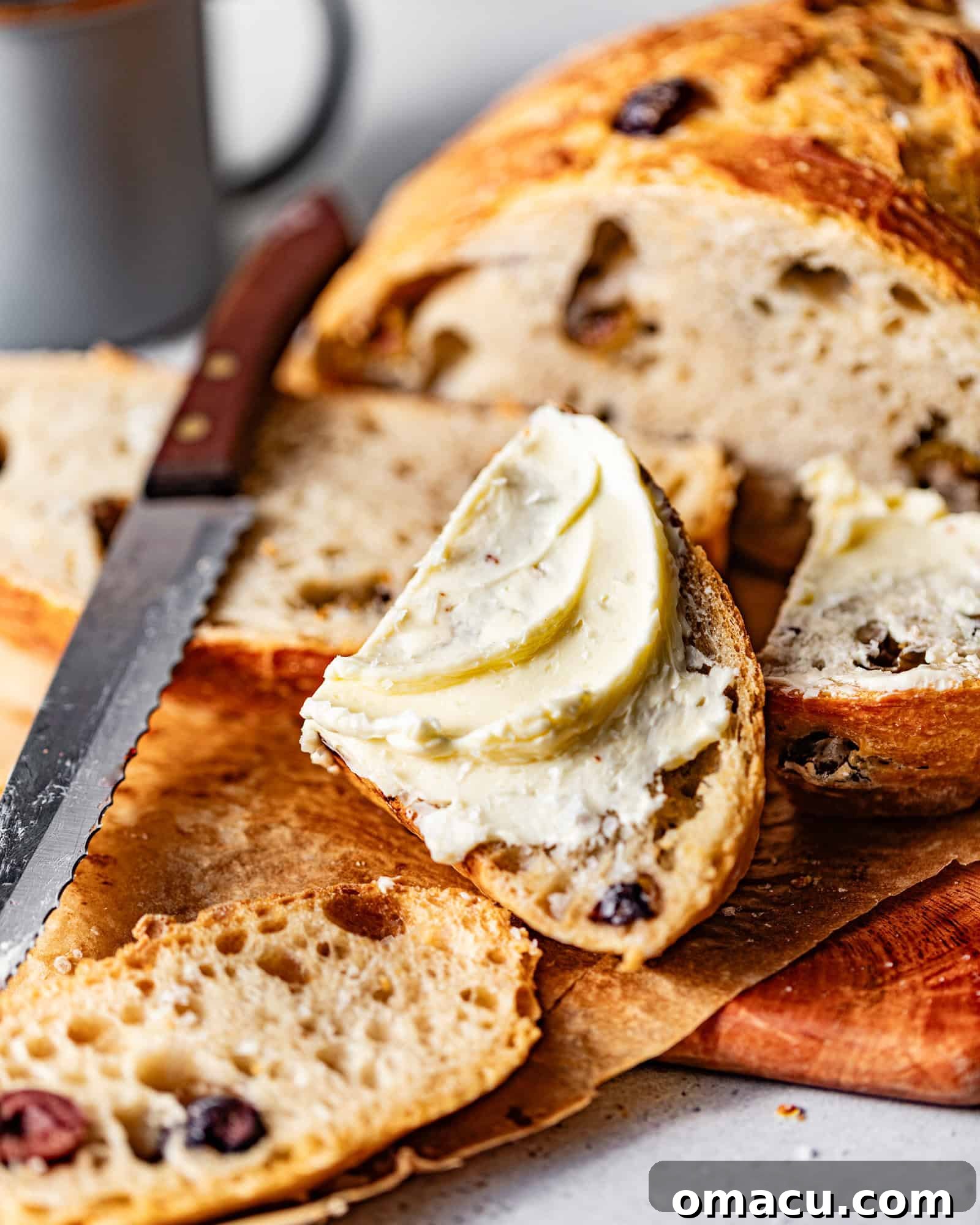 A perfectly sliced no-knead olive bread on a cutting board, with a bread knife and a small piece of bread topped with butter.