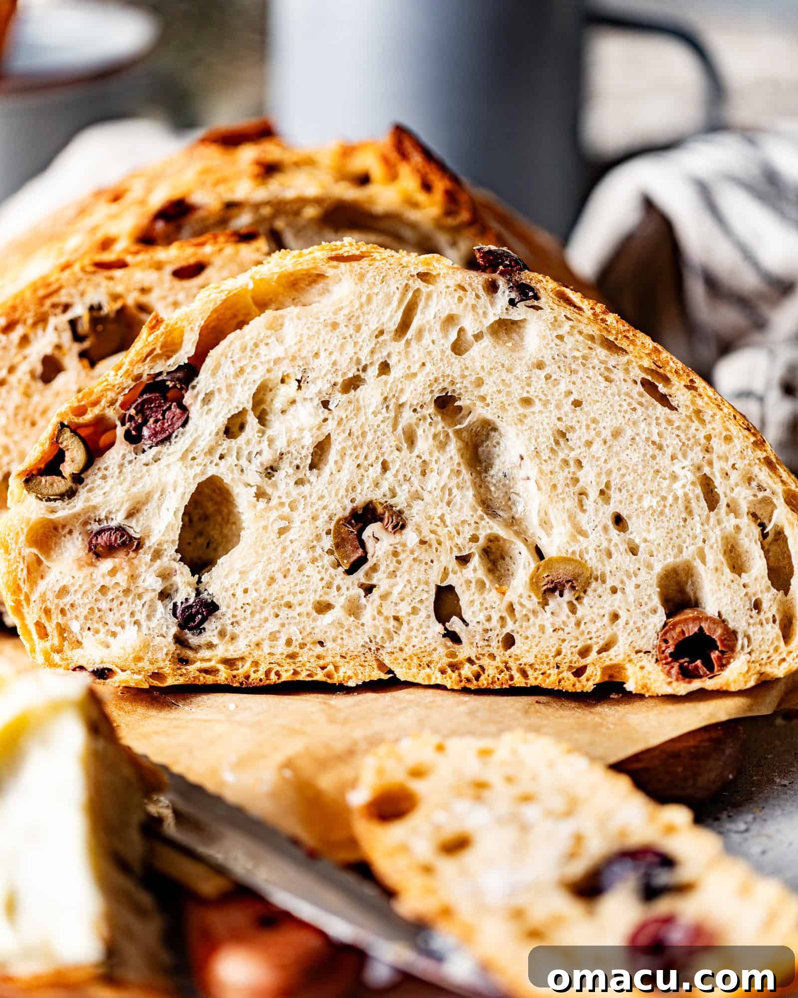 A freshly baked no-knead olive bread on a brown cutting board, next to a blue mug and a bread knife.