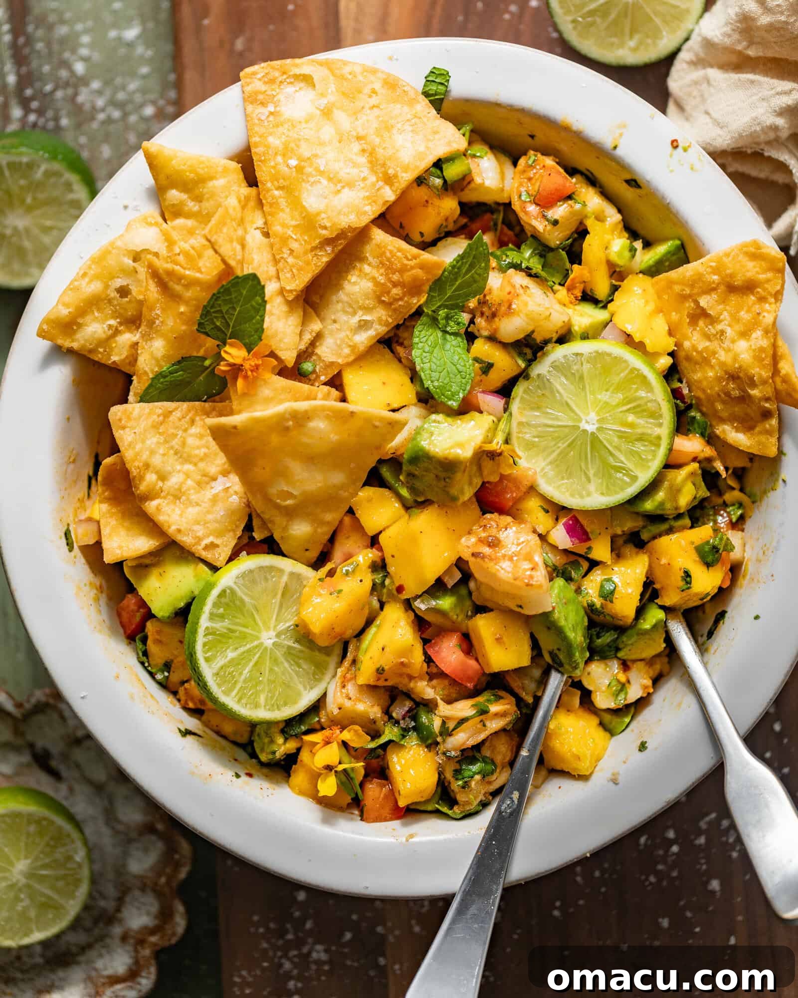 Vibrant mango shrimp salad in a white bowl, garnished with crispy tortilla chips, fresh lime wedges, and delicate edible flowers.