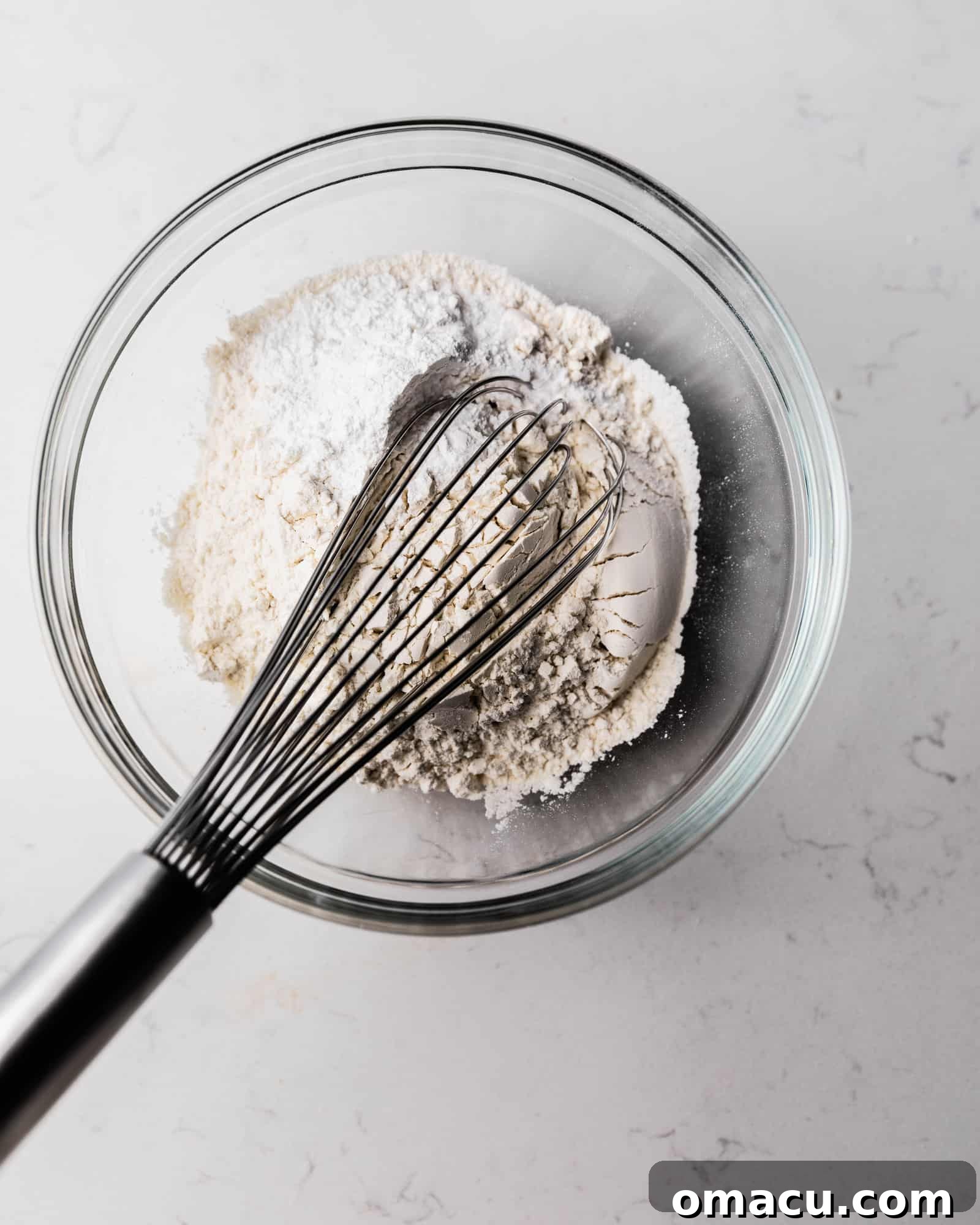 A bowl of dry ingredients, including flour, baking powder, baking soda, and salt, with a whisk ready to combine them evenly.
