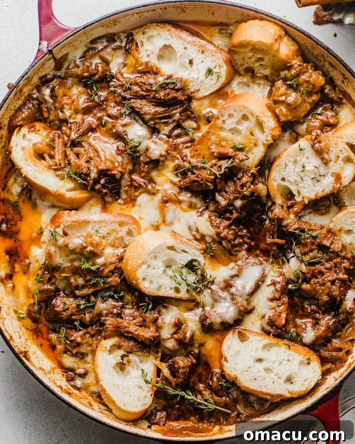 overhead photo of french onion pot roast in a braising pan topped with gruyere cheese, fresh thyme, and sliced baguettes.