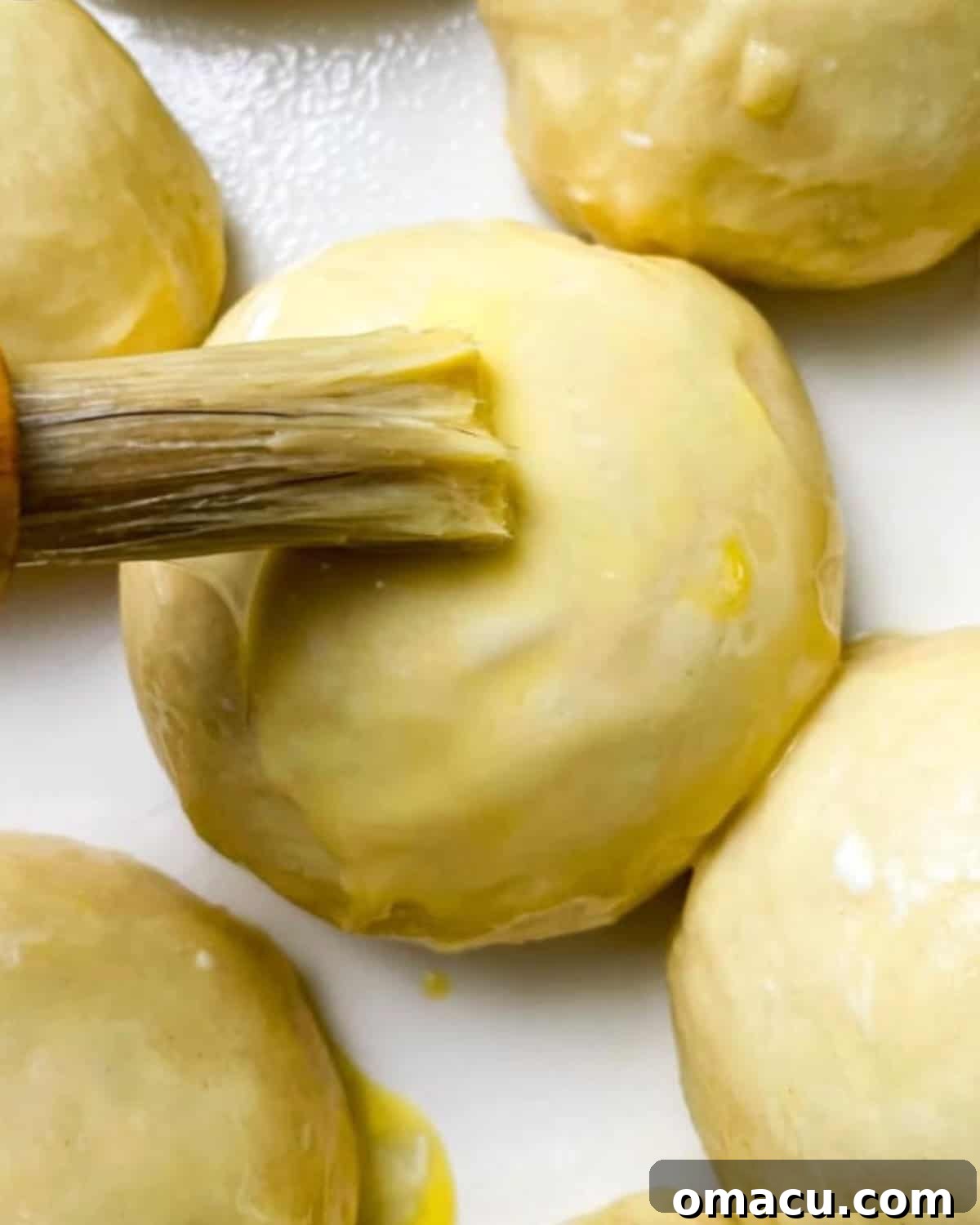 A pastry brush applying a thin layer of egg wash to the tops of unbaked Hawaiian rolls in a baking dish.