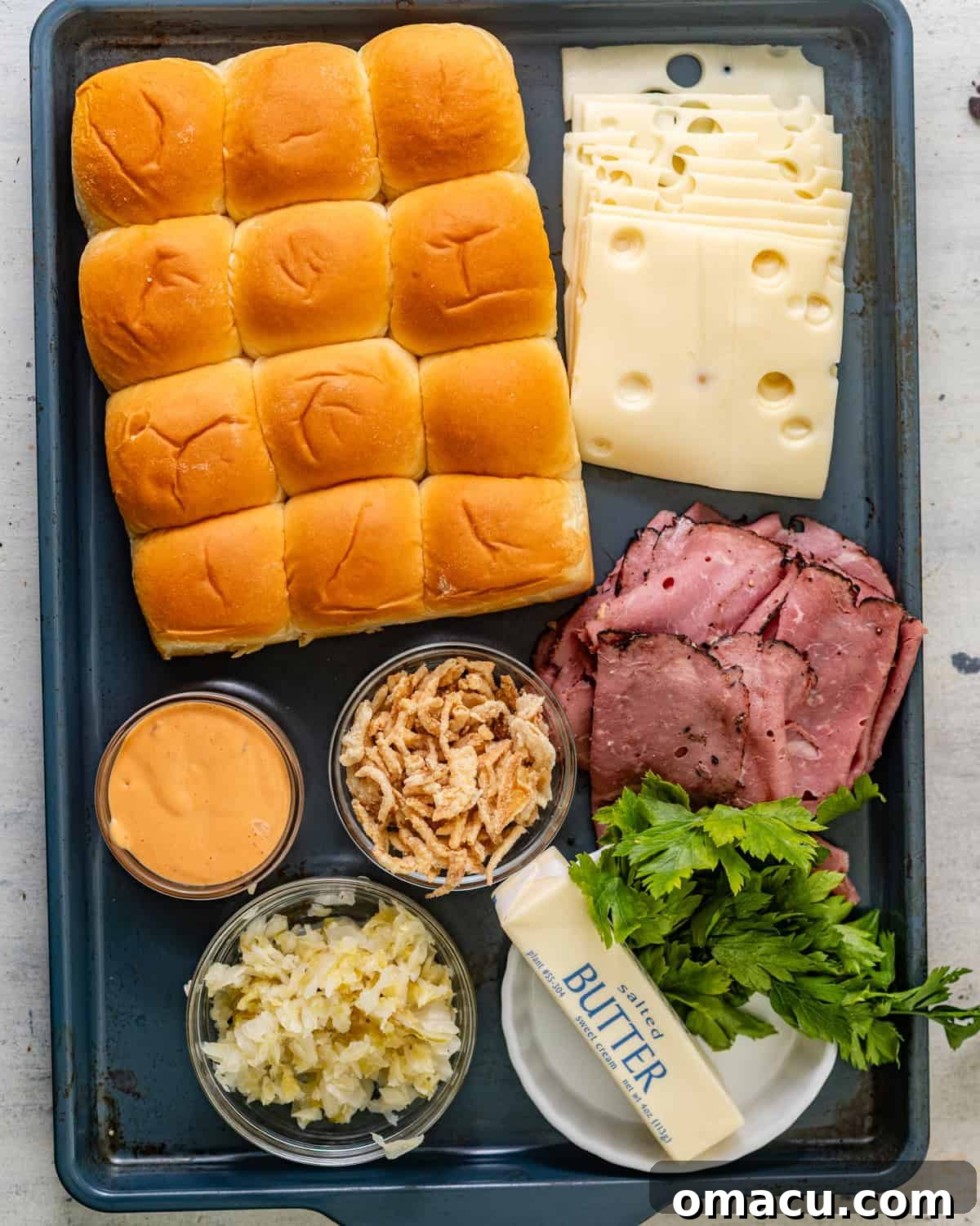 A beautifully arranged flat lay of all the fresh ingredients required to prepare delightful Reuben sliders: a package of Hawaiian rolls, slices of Swiss cheese, pastrami, a block of butter, a container of crispy French fried onions, a bottle of Thousand Island dressing, a jar of sauerkraut, fresh garlic cloves, chopped parsley, and poppy seeds.