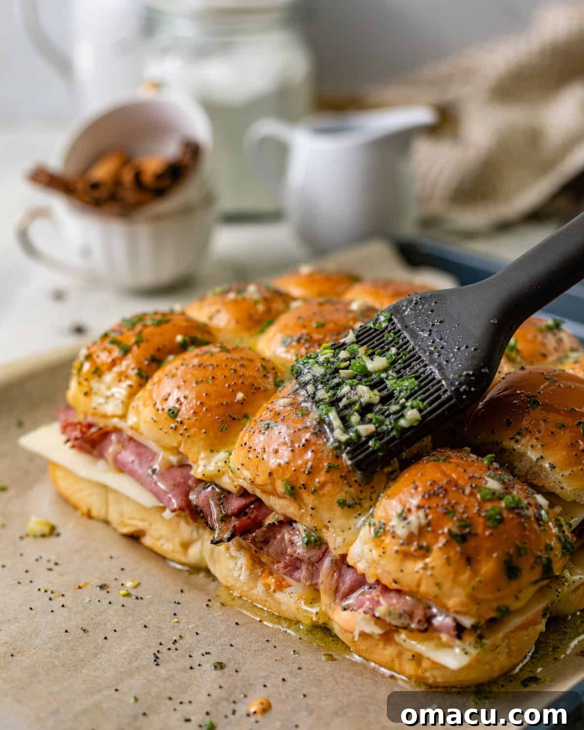 A pastry brush skillfully applying a savory mixture of garlic butter and poppy seeds onto the top buns of the assembled Reuben sliders, giving them a glossy finish.