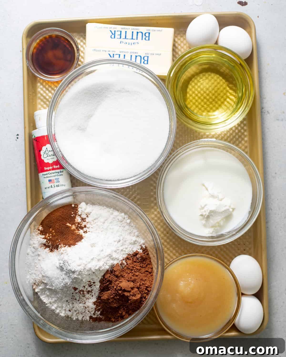 A beautiful flat lay of all the essential red velvet cake ingredients laid out on a baking tray, including flour, cocoa powder, baking powder, baking soda, salt, milk powder, oil, butter, sugar, brown sugar, eggs, vibrant red gel food coloring, espresso powder, buttermilk, sour cream, and hot coffee.