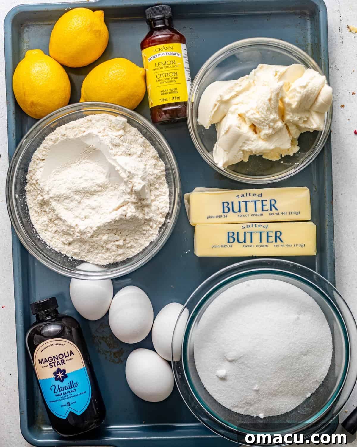 A collection of fresh ingredients laid out on a baking tray, including sticks of butter, granulated sugar, whole lemons, eggs, bottles of vanilla, lemon, and almond extract, whole milk, and a tub of ricotta cheese.