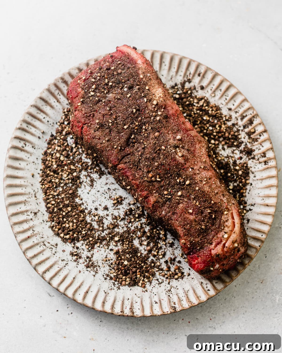 New York strip steak on a plate covered in crushed peppercorns, ready for searing.