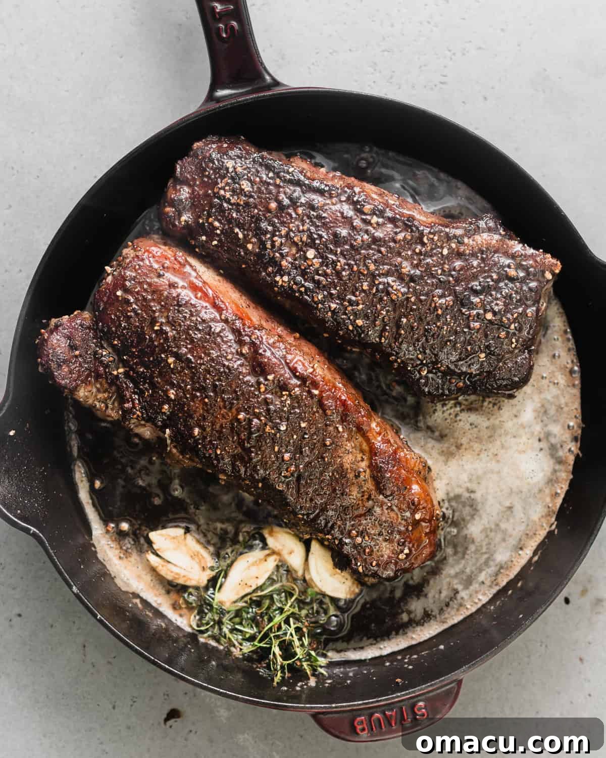 Seared steaks in a cast iron skillet with butter, smashed garlic, and sprigs of thyme and rosemary, being basted.