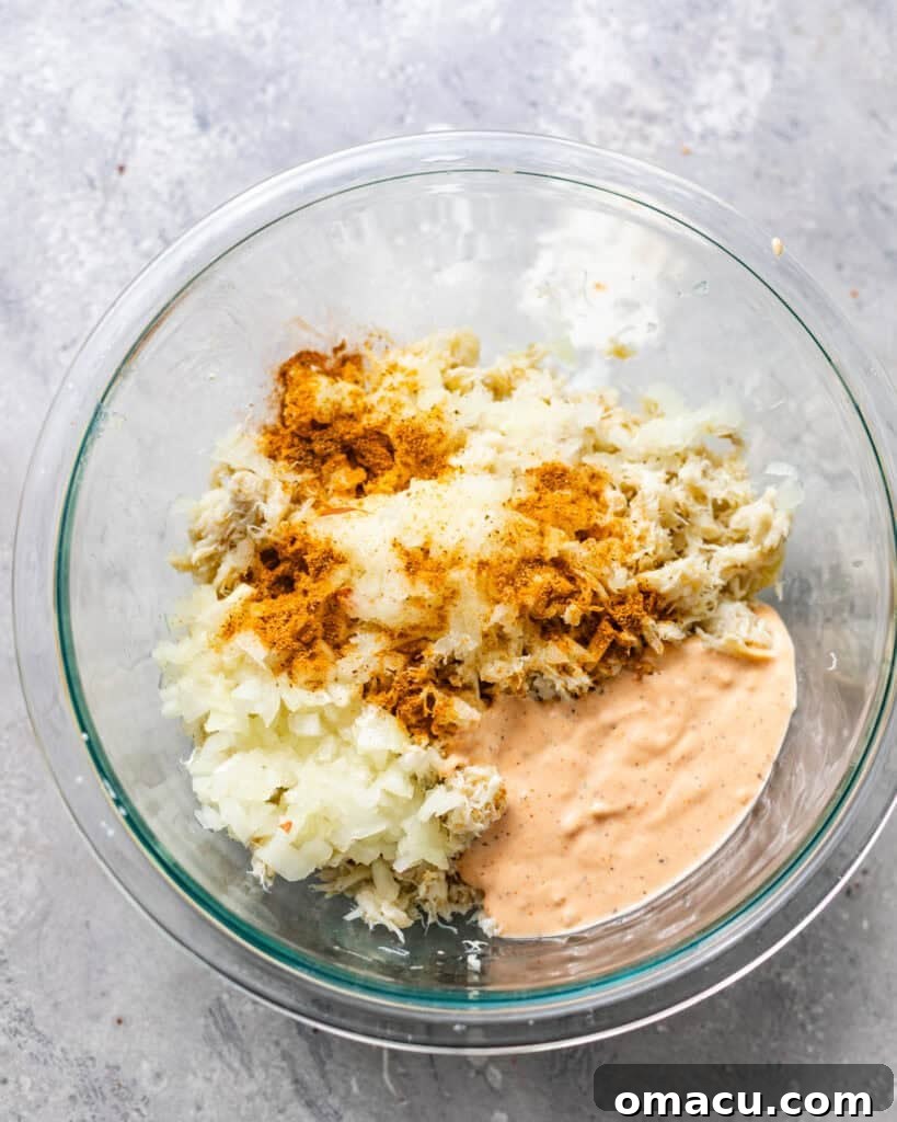 A mixing bowl containing lump crab meat, seasonings, a portion of remoulade sauce, whisked eggs, grated onion, and milk, ready to be combined.