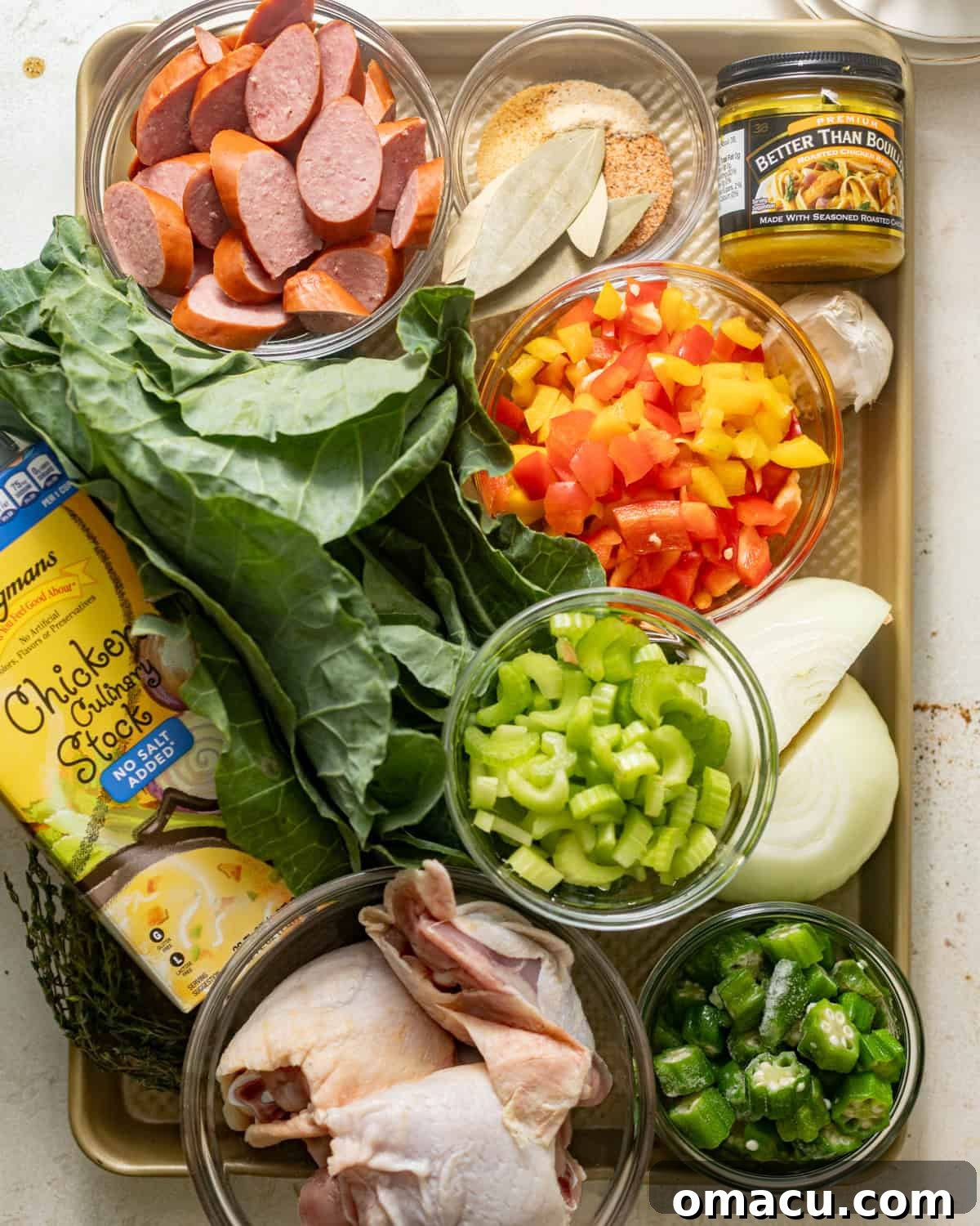 A selection of fresh ingredients laid out on a baking tray, including vibrant collard greens, colorful bell peppers, onion, celery, slices of sausage, raw chicken thighs, chicken bouillon, chicken stock, various seasonings, okra, Worcestershire sauce, file powder, and fresh garlic.