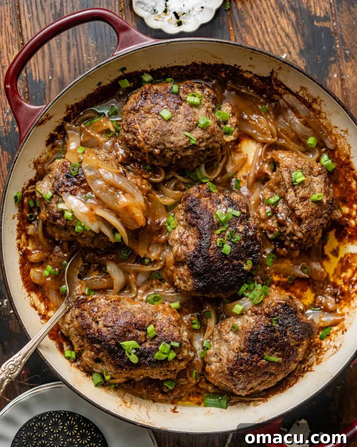 Overhead photo of three golden-brown baked hamburger steaks swimming in a rich, dark brown onion gravy, served in a cast-iron skillet.