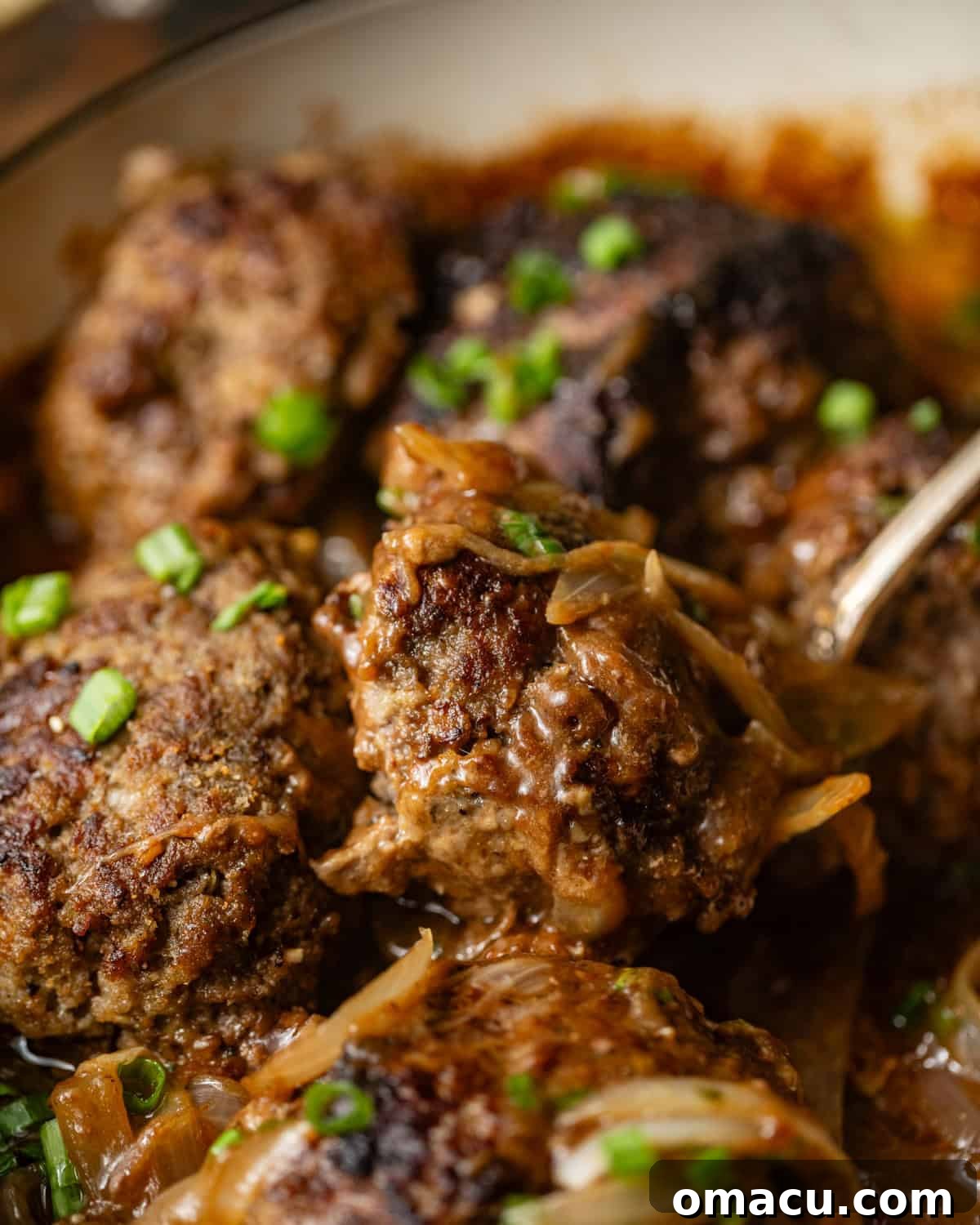 Close-up photo of a serving of baked hamburger steaks with onions and gravy in a cast-iron skillet, with a large serving spoon.