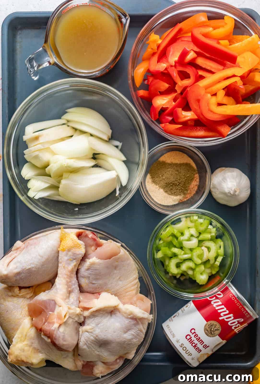 ingredients to make smothered chicken on a baking tray - chicken legs, bell peppers, onions, celery, cream of chicken soup, seasonings, garlic, and chicken broth.