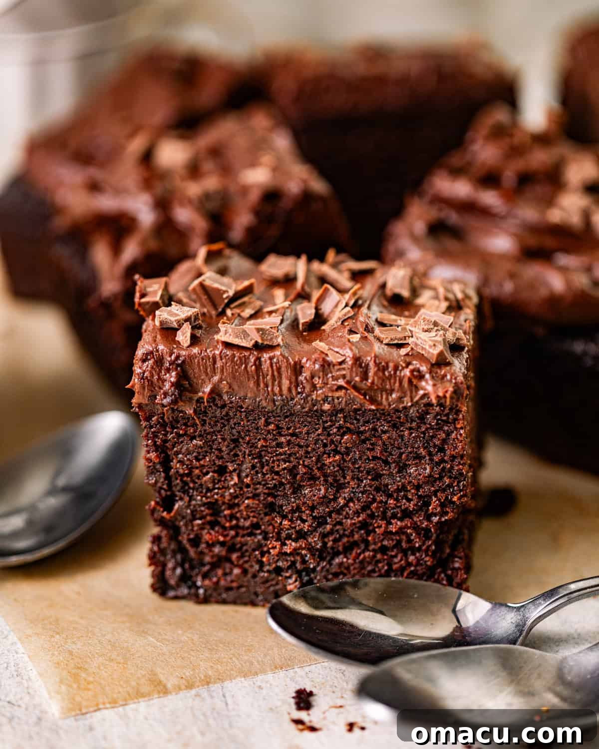 A delectable slice of chocolate snack cake, generously topped with shimmering chocolate ganache and delicate chocolate shavings. The cake sits elegantly on a table beside a spoon, inviting a delightful bite.
