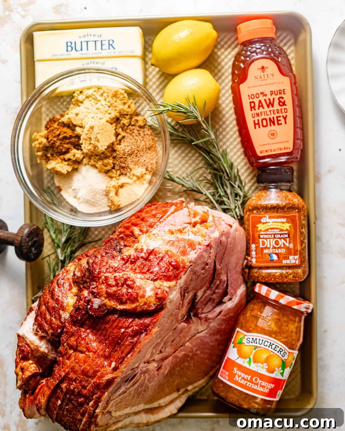 All the fresh ingredients laid out on a baking sheet: a bone-in spiral ham, brown sugar, Dijon mustard, melted butter, honey, vibrant orange marmalade, fresh lemons, pineapple juice, and a selection of aromatic seasonings.