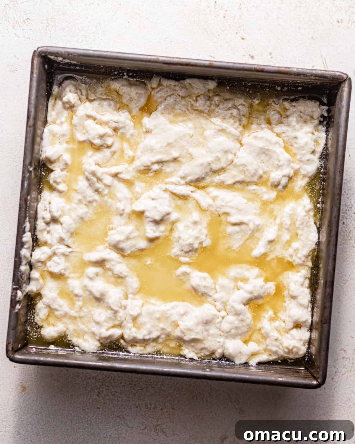 Biscuit dough carefully spread evenly in a square baking pan, sitting glistening in a pool of melted butter.