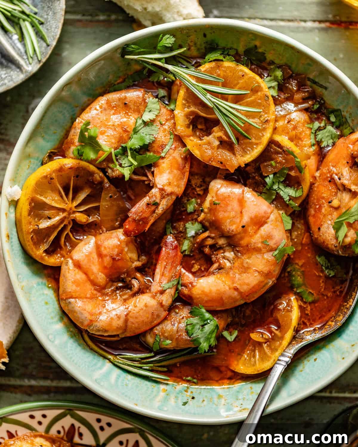 new orleans bbq shrimp in a bowl with a spoon garnished with fresh parsley and rosemary alongside a piece of crusty bread.