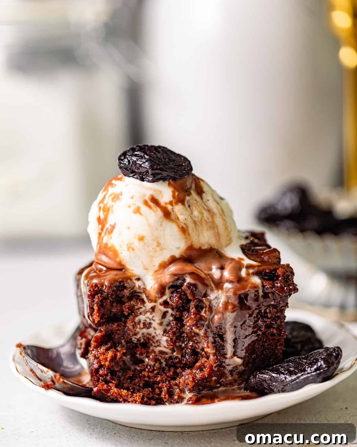 a slice of chocolate sticky toffee pudding on a small plate topped with ice cream and a prune.