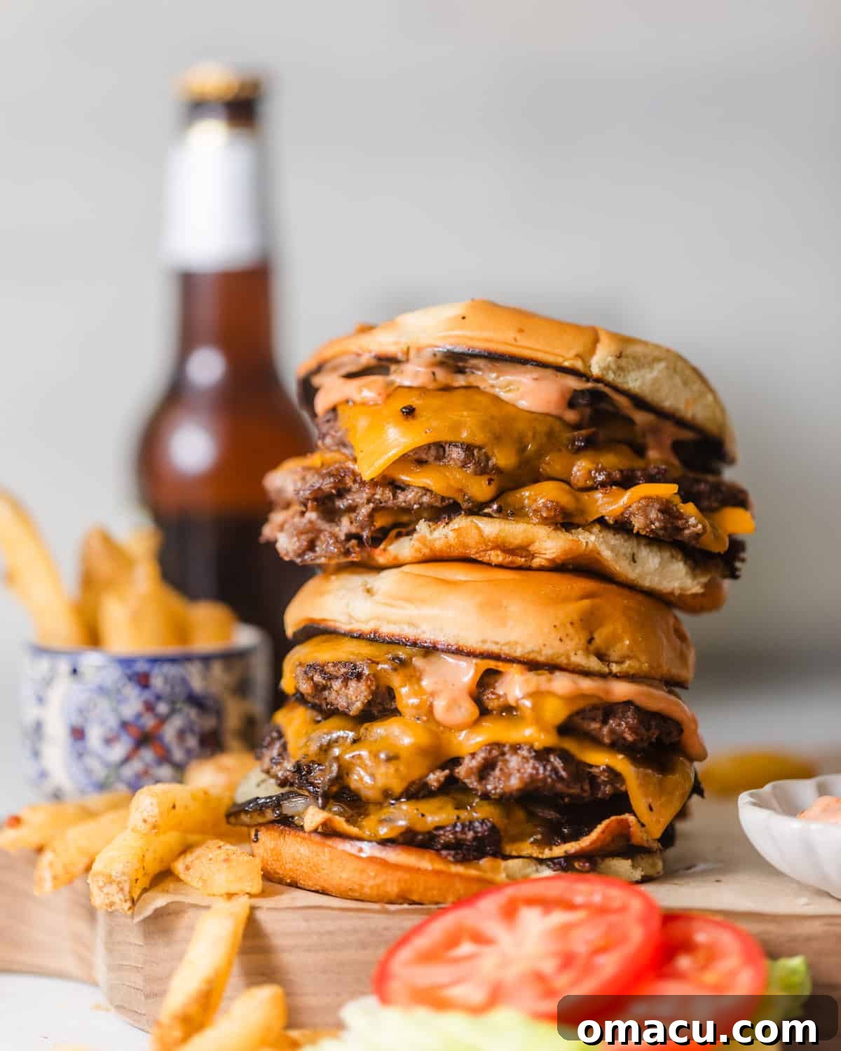 smash burgers stacked on top of each other on a serving board with french fries, lettuce, and tomato on the side.