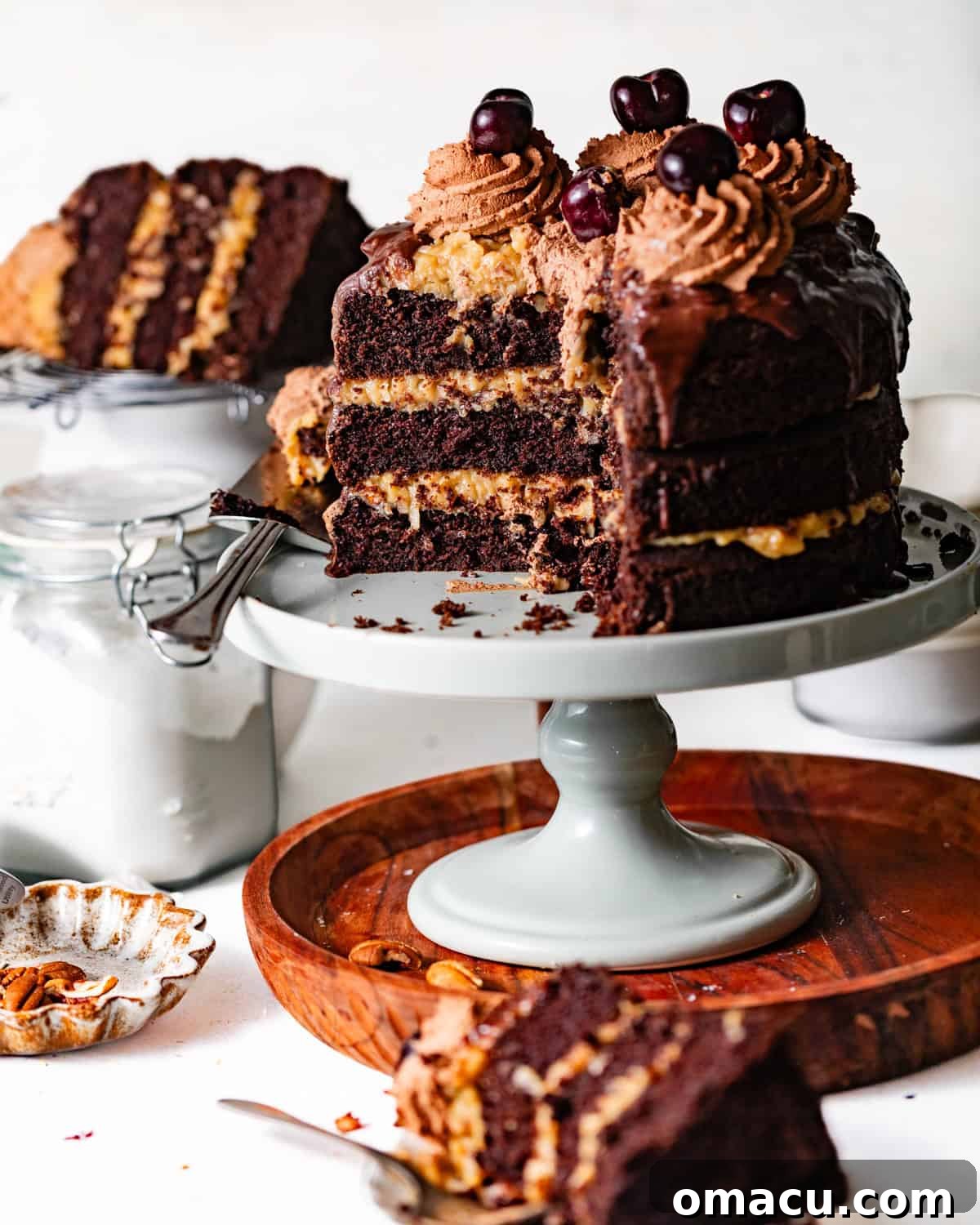 German chocolate cake on a cake stand topped with chocolate ganache and chocolate whipped cream and cherries for garnish.