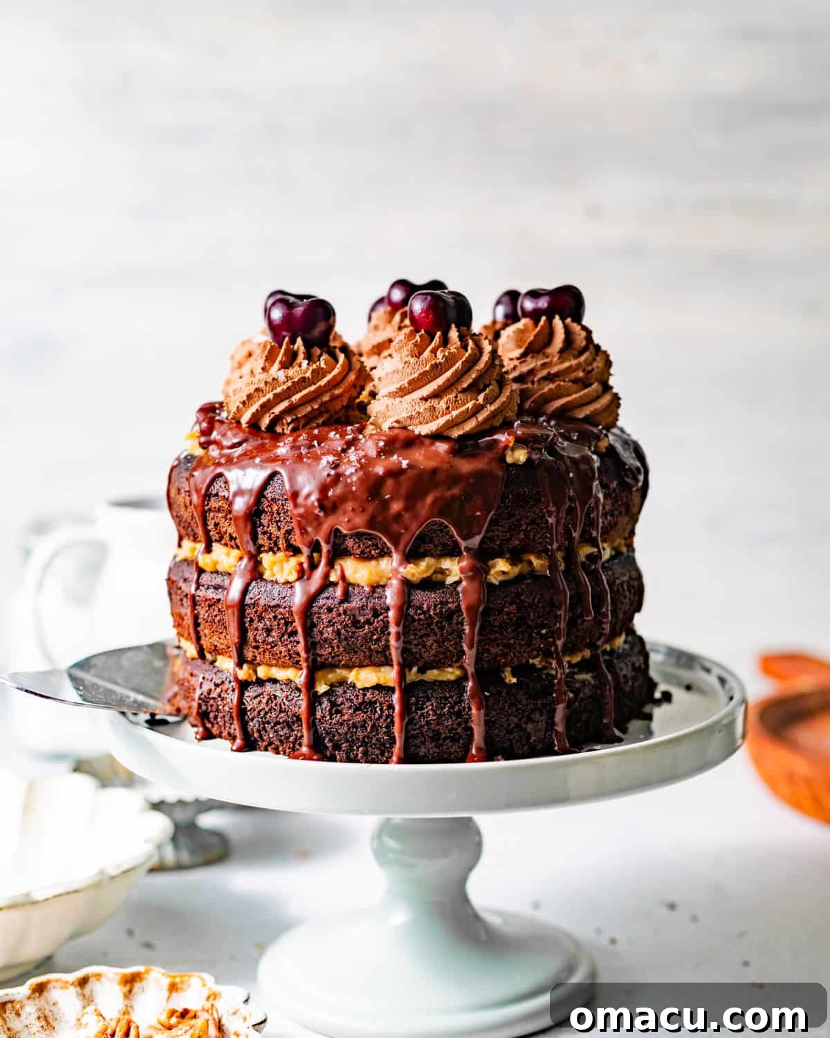 german chocolate cake on a cake stand with chocolate ganache and chocolate whipped cream and cherries on top.