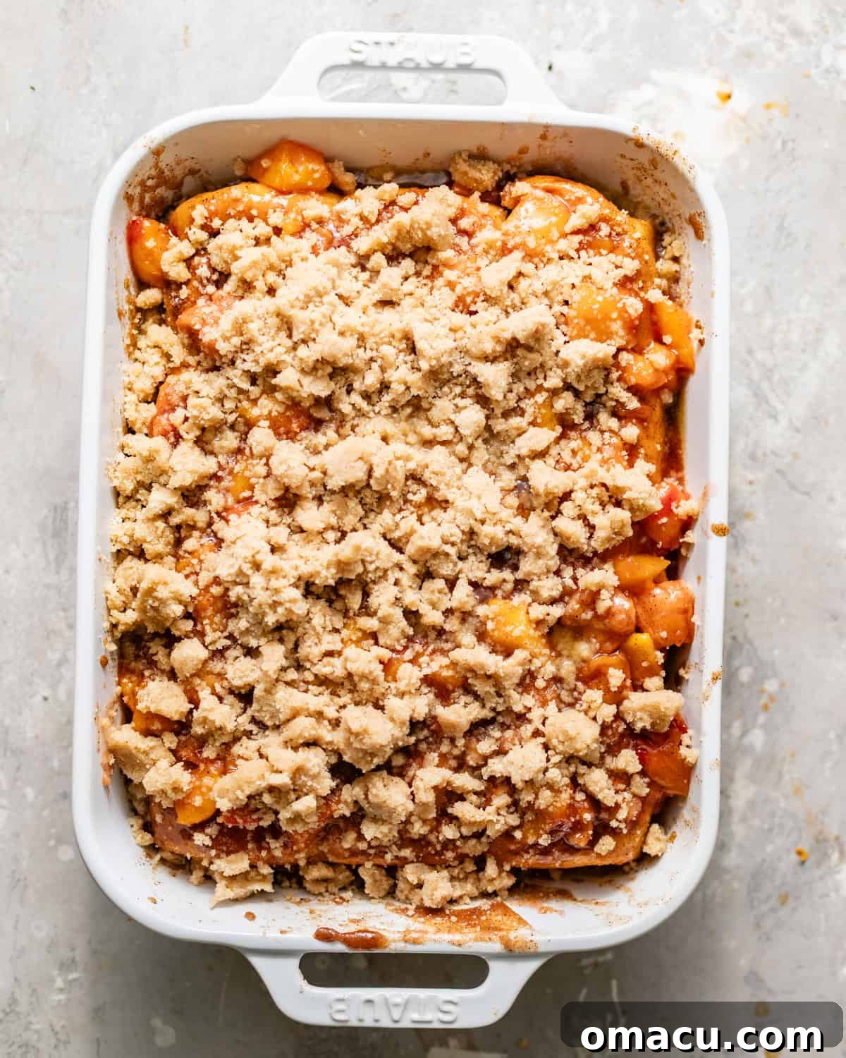 peach cobbler cinnamon roll bites assembled in a baking dish - hawaiian rolls covered with cinnamon roll filling and filled with peach cobbler and topped with streusel.