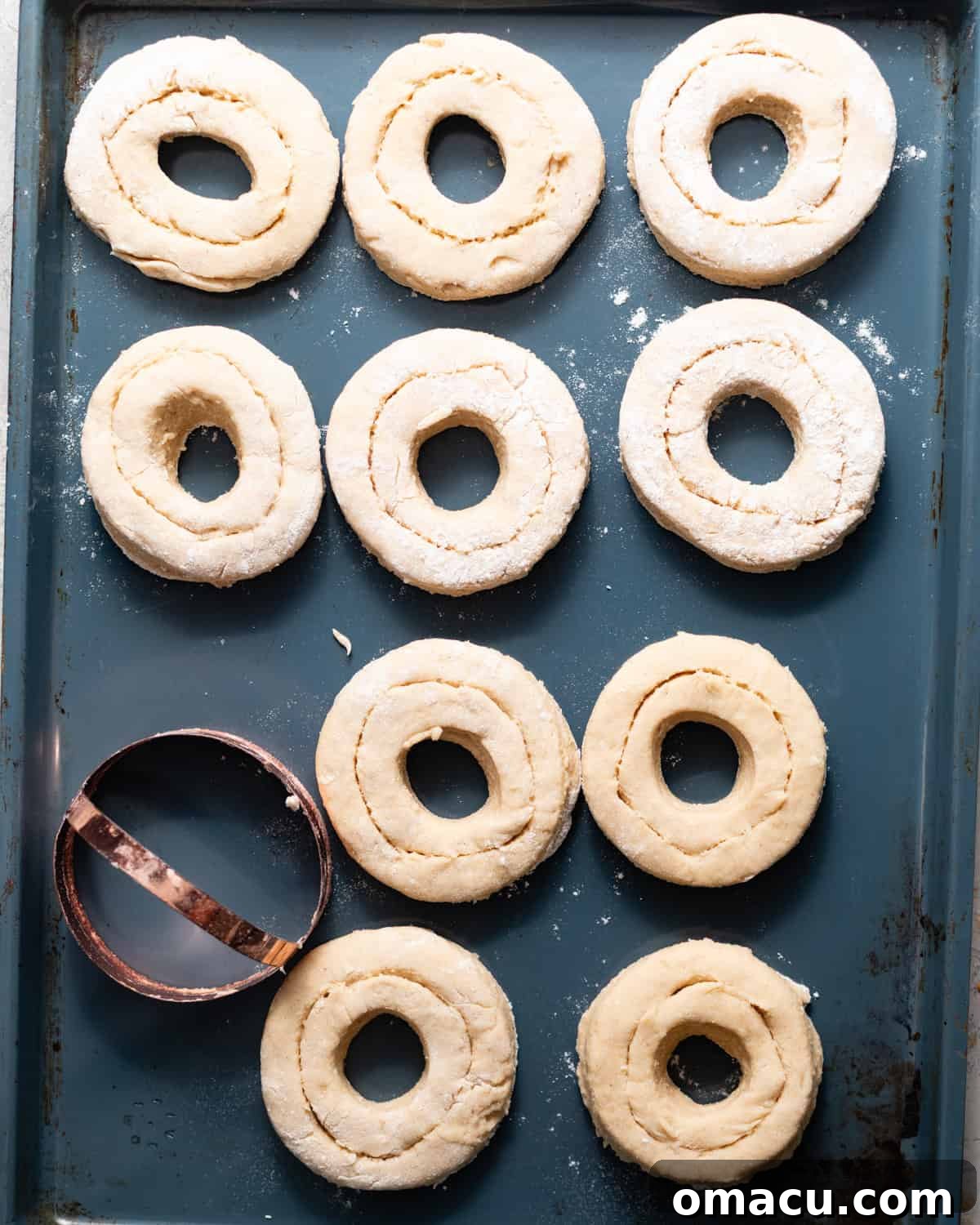 Classic Maple Cake Donuts Recipe 6 Unfried maple cake donuts with scored tops arranged on a baking sheet beside a round donut cutter.