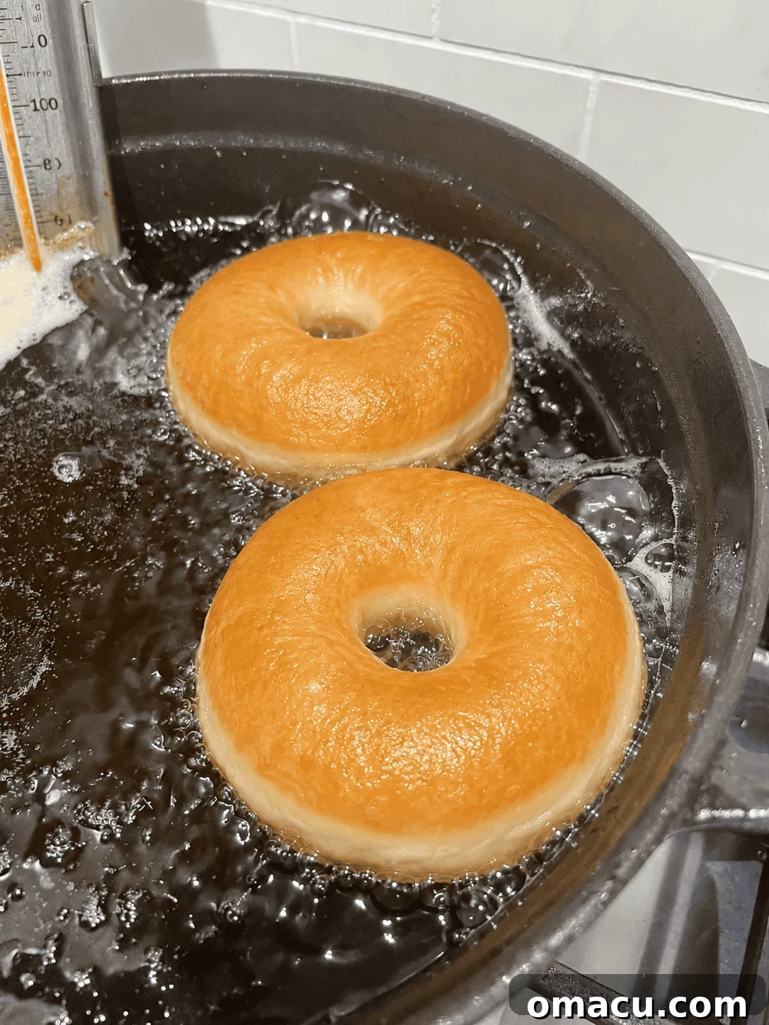 Two golden brown yeast donuts frying in bubbling oil with a candy thermometer in a dutch oven.