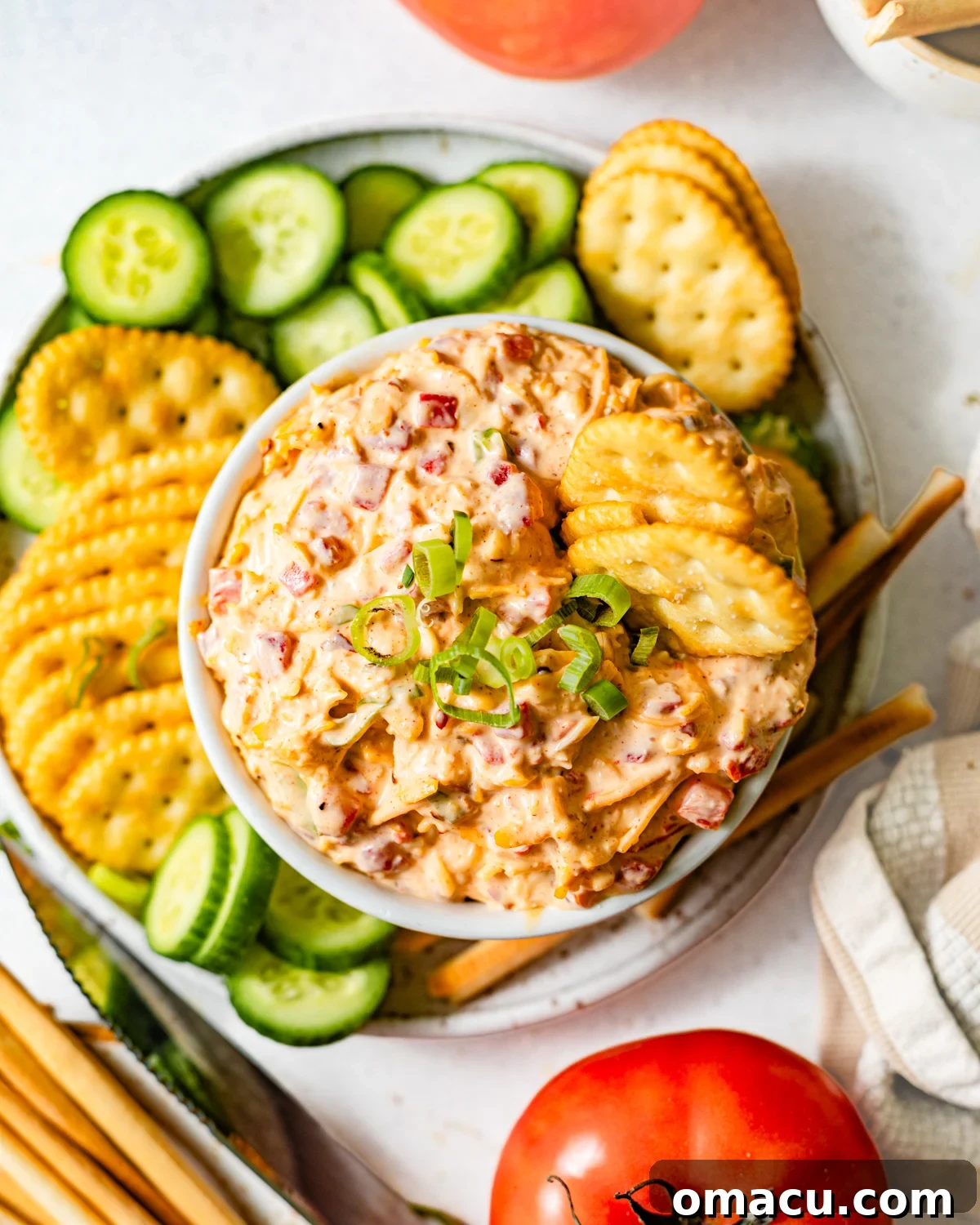 Creamy Southern Pimento Cheese Dip for Game Day 2 pimento cheese in a bowl next to sliced cucumbers, tomatoes, crackers, and breadsticks for dipping.