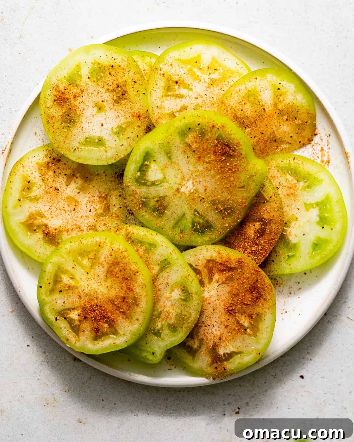 Crispy Southern Fried Green Tomatoes Recipe 4 sliced green tomatoes on a plate sprinkled with Cajun seasoning.