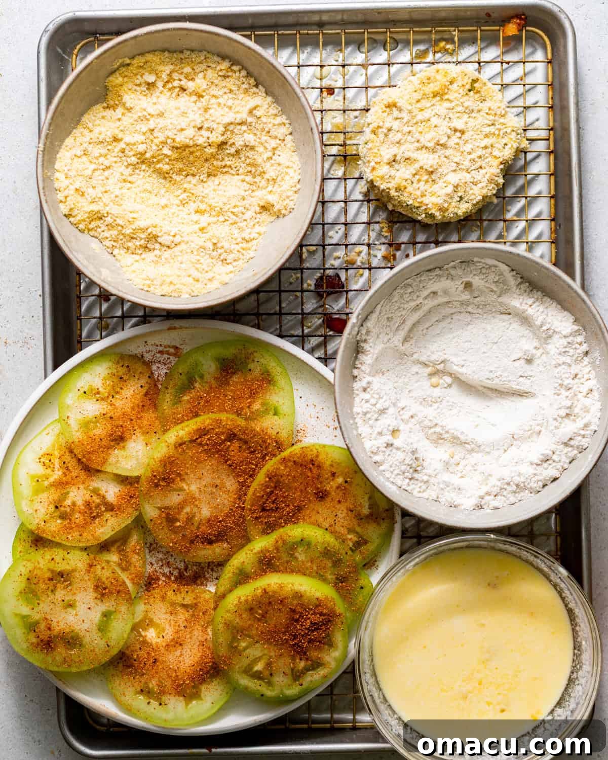 Crispy Southern Fried Green Tomatoes Recipe 5 breading station set up on a baking sheet - a bowl with buttermilk and eggs whisked together, another bowl with just flour, and another bowl with cornmeal and breadcrumbs mixed together.
