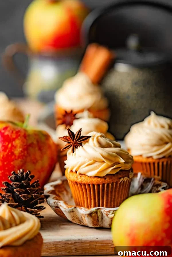 apple cider cupcakes filled with apple butter topped with cinnamon cream cheese frosting surrounded by apples on a cutting board.
